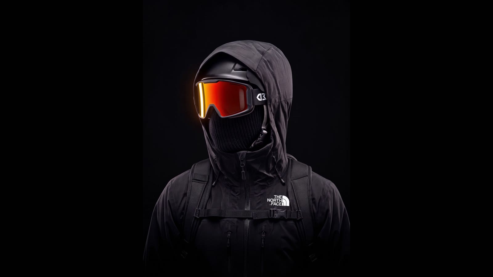 Hooded figure in a black shell jacket with orange ski goggles, on black